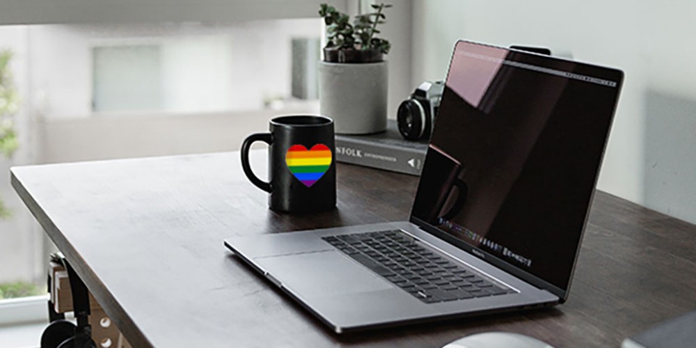Office table: open laptop, black mug with heart-shaped LGBT flag, symbolizing care for returning LGBT employees